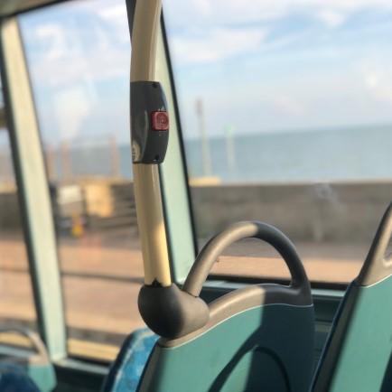 A bell on a support pole on a bus. A seafront can be seen outside of the window in the background