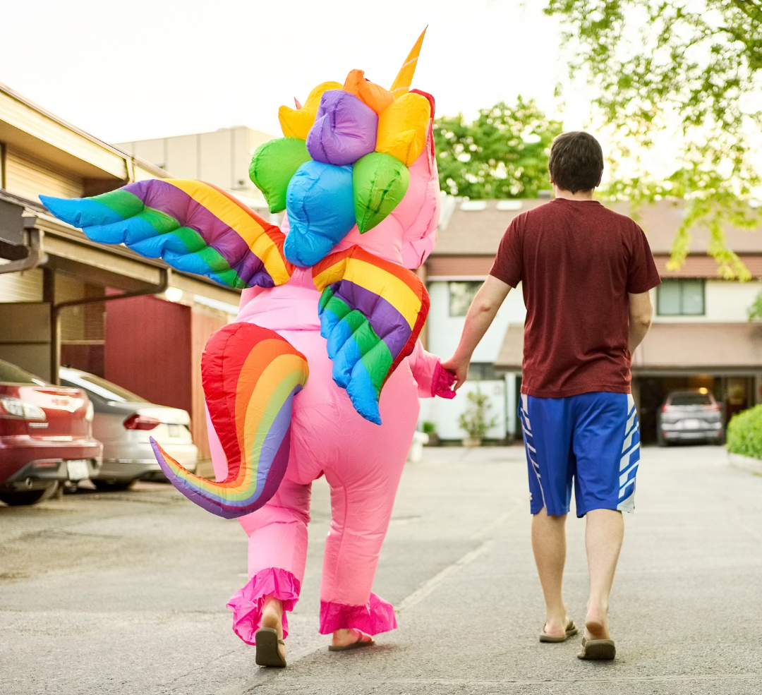 A person dressed up as a colourful unicorn walking away from the camera. A man is holding hands with the unicorn.