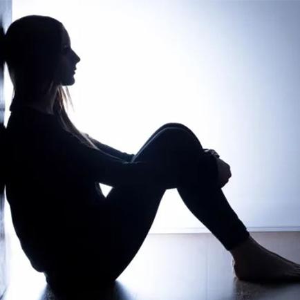 A silhouette of a woman sitting on the floor with her back on a wall and her arms wrapped around her knees