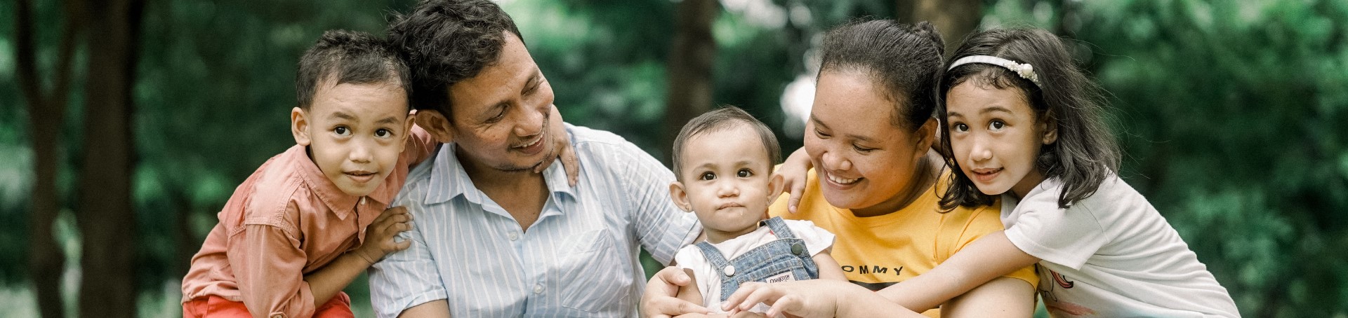A family of Asian origin, there are of five of them sitting outdoors. The mother and father are carrying a toddler and there are other two small children next to each of them.