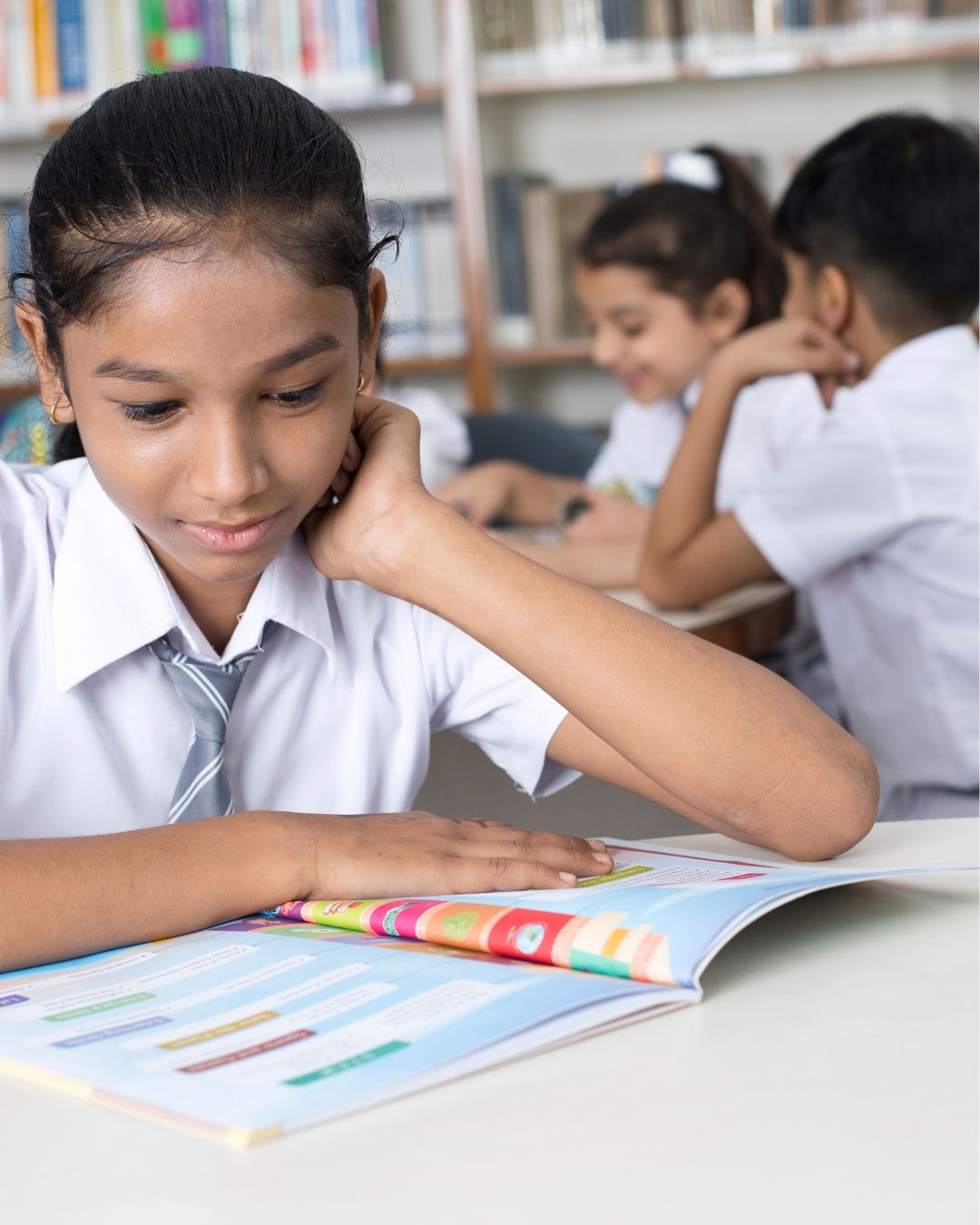 Photograph of 3 children in a classroom. They are all wearing school shirts and ties. In the background a girl and boy sit at a table. In the foreground a girl aged around 11 with her hair in a pony tail is sat at a table looking at an open book.