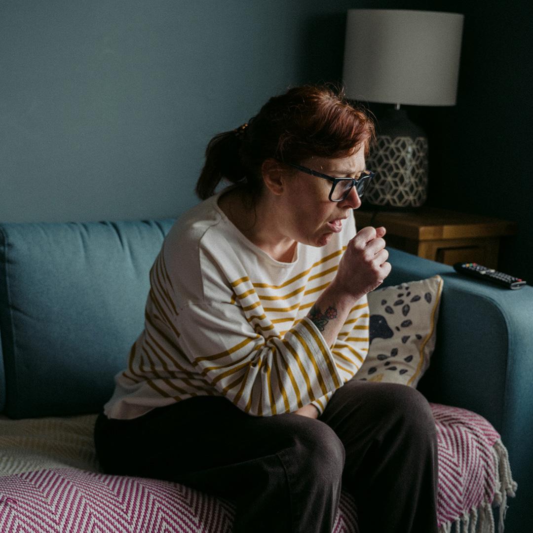 A middle-aged woman sitting on a sofa alone, coughing into her hand