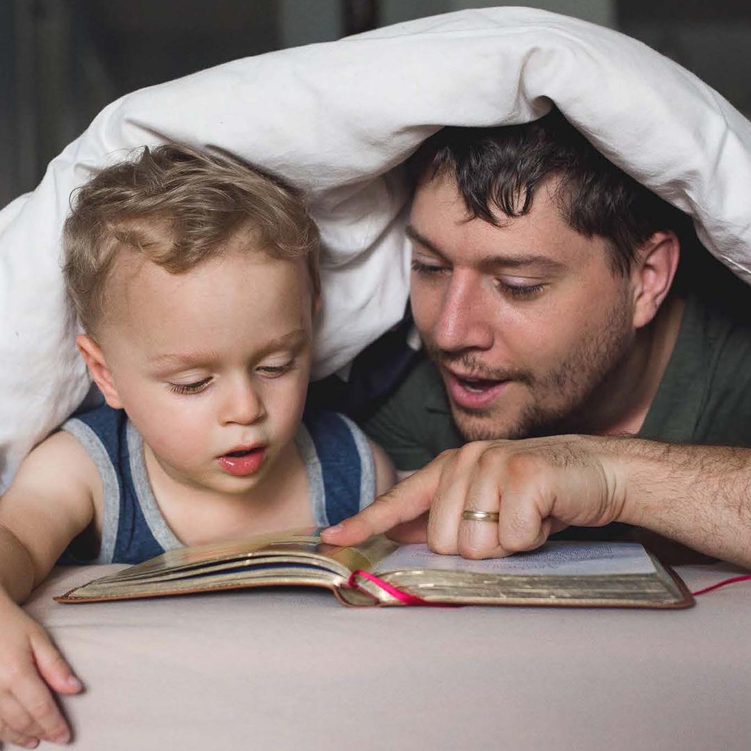 Father and child about 2 years old reading a book together under a duvet