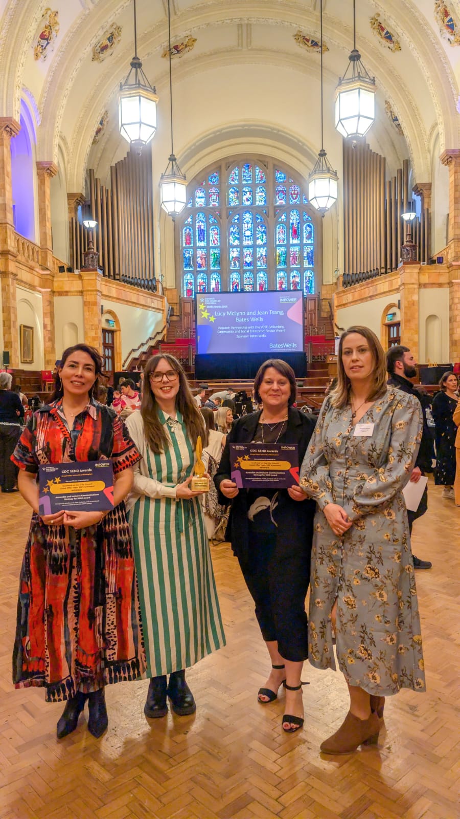 a group of 4 adults stand in a large grand hall with a stained glass window in the background. One of the group is holding a trophy.