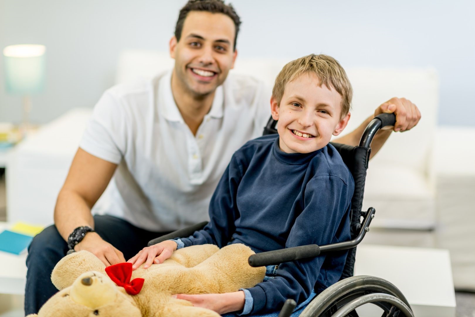 A photograph of a dark haired man crouched down next to a blonde haired boy in a wheelchair. They are both smiling and the boy is holding a teddy bear.