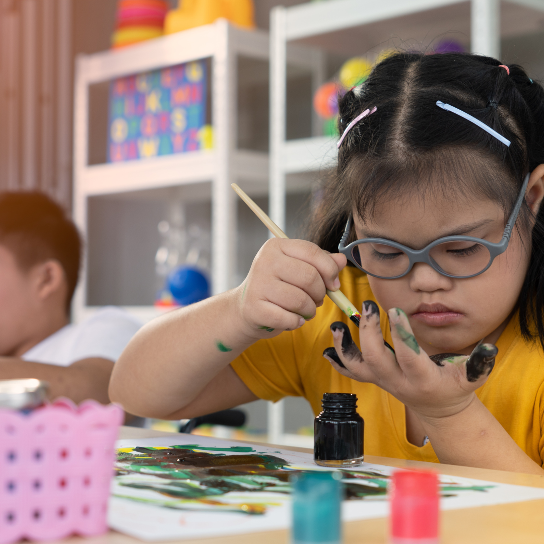 photograph of a young girl with glasses sat at a desk in a colourful classroom. She is painting her fingers with a paintbrush.
