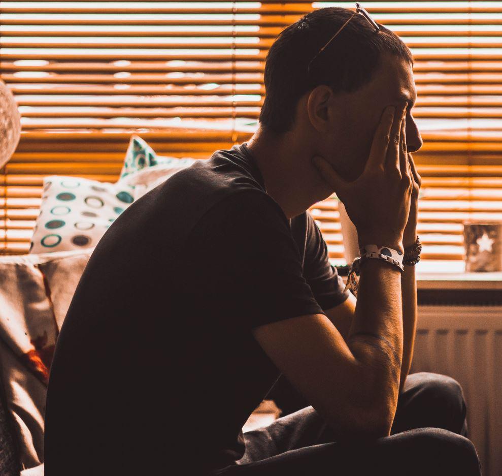 Silhouette of man sitting, with his head in his hands, in front of a blind. The suggestion is the man is struggling mentally