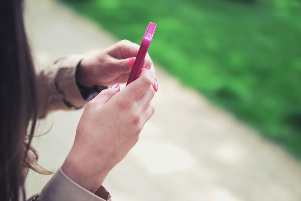 Woman's hands holding a pink smartphone with both hands in a casual outdoor setting.