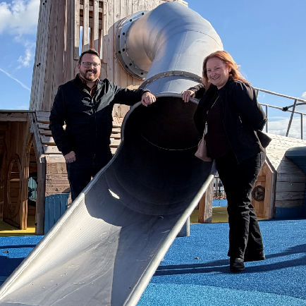 Photograph of 2 smiling adults standing either side of a tube slide in a playground.